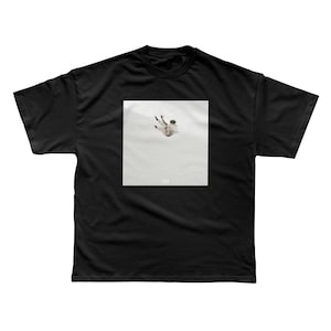 May include: Black t-shirt featuring a square graphic of a person falling. The graphic is in shades of white and beige, set against a light background. The t-shirt is made of a soft, comfortable material.
