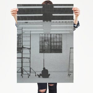 May include: Black and white print of a person sitting on the floor in front of a window. The print has a minimalist aesthetic, with a focus on the silhouette of the person and the industrial setting.