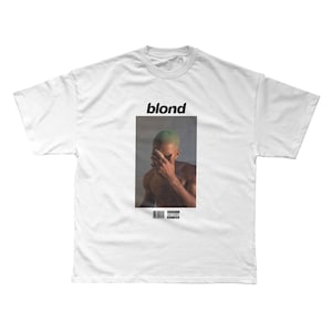 May include: White t-shirt featuring the word "blond" above a rectangular image. The image shows a person with green hair covering their face with their hand. A barcode and advisory warning are below the image.