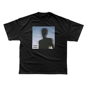 May include: Black t-shirt featuring a blurred silhouette of a person against a blue and white background. The shirt has a square graphic with a parental advisory warning. A casual, unisex tee.