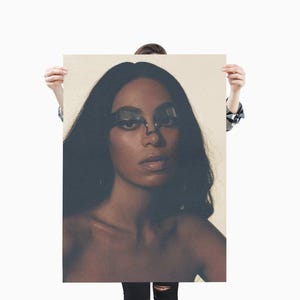 May include: A portrait of a person with long, dark hair and a neutral expression. They are wearing unique, dark-colored eyewear. The image is held up by a person, with a neutral background.