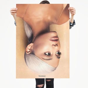 Puede incluir: Un póster con un retrato de una persona con piel clara y cabello blanco. La persona está mirando hacia arriba, con la palabra "Sweetener" impresa en la parte inferior. El póster está sostenido por una persona que lleva una camisa de mezclilla y pantalones negros.