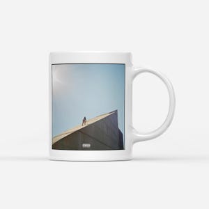 May include: White ceramic mug featuring a photograph of a person standing on a building's edge against a blue sky. The image has a vintage aesthetic, with a parental advisory warning at the bottom.