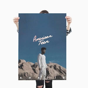 May include: A poster with the words "American Teen" in a cursive font against a blue sky background. The poster features a person wearing white clothing standing in front of a rocky landscape. The name "Khalid" is printed at the bottom.