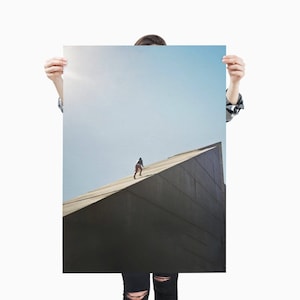 May include: A photographic print featuring a person walking on a large, angular, dark gray structure against a light blue sky. The person is small in the frame, adding a sense of scale and adventure. The print is held up by a person.
