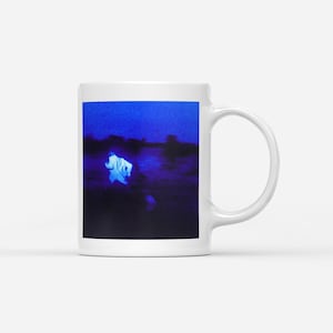 May include: White ceramic mug featuring a surreal image of a figure in a blue landscape. The artwork has a dark, moody aesthetic with a focus on the contrast between light and shadow. The mug has a standard handle and a glossy finish.