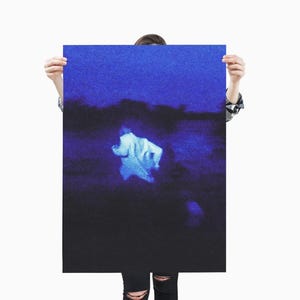 May include: A poster with a dark blue and black color scheme. The image features a blurry, light-colored figure against a dark background. The poster is held up by a person wearing a black and white plaid shirt and black jeans.