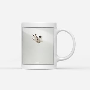 May include: White ceramic mug featuring a framed image of a person in mid-air against a light background. The person is wearing light-colored clothing. The mug has a curved handle and a simple, clean design.
