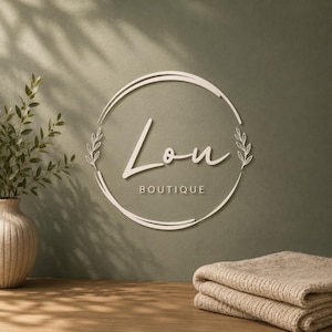 Può includere: Un'insegna da parete circolare con il testo Lou BOUTIQUE in un carattere corsivo e sans-serif, racchiuso da un cerchio a linea sottile con accenti di foglie sui lati. L'insegna è montata su una parete verde oliva con ombre soffuse.