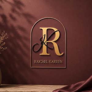 Può includere: Un'insegna da parete con una cornice ad arco color oro, una grande lettera R dorata e una lettera K nera in corsivo. Il testo RAICHEL KARREN è stampato in oro sotto le lettere. L'insegna è esposta su una parete color bordeaux scuro.