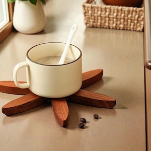 May include: A cream-colored mug with a dark brown rim and handle, holding a light-colored liquid and a white spoon. The mug rests on a dark brown, wooden, flower-shaped coaster. Scattered around are a few coffee beans. The scene suggests a cozy coffee or tea break.
