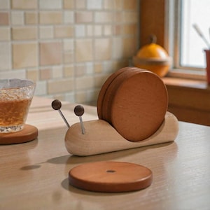 May include: A wooden snail-shaped coaster holder with a set of brown coasters. The holder is light-colored wood, and the coasters are a darker brown. The snail has two antennae with small, round tips.