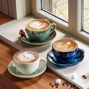 Könnte beinhalten: Drei Keramik-Kaffeetassen mit Latte Art, jede auf einer passenden Untertasse. Die Tassen sind in Grün-, Blau- und Weißtönen gehalten, wobei die Latte Art ein Herz, einen Farn und ein Bärengesicht zeigt. Die Tassen stehen auf einer Holzoberfläche.