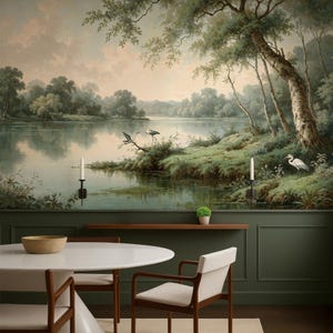 Puede incluir: Un mural de pared con una pintura de paisaje de un lago tranquilo con garzas, árboles y un cielo suave. Debajo del mural hay una pared con paneles de color verde oscuro, un estante de madera con una planta pequeña, una mesa redonda blanca y dos sillas.