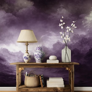 May include: A wooden console table featuring a beige lampshade, a blue and white porcelain jar, a clear glass vase with white flowers, and stacks of white books. A woven basket holds books on the lower shelf against a purple cloudy mural.