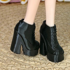 May include: A pair of black doll-sized ankle boots featuring a thick platform sole and a chunky block heel. The boots have a lace-up front design with visible horizontal texture lines across the surface, extending up to the ankle area.