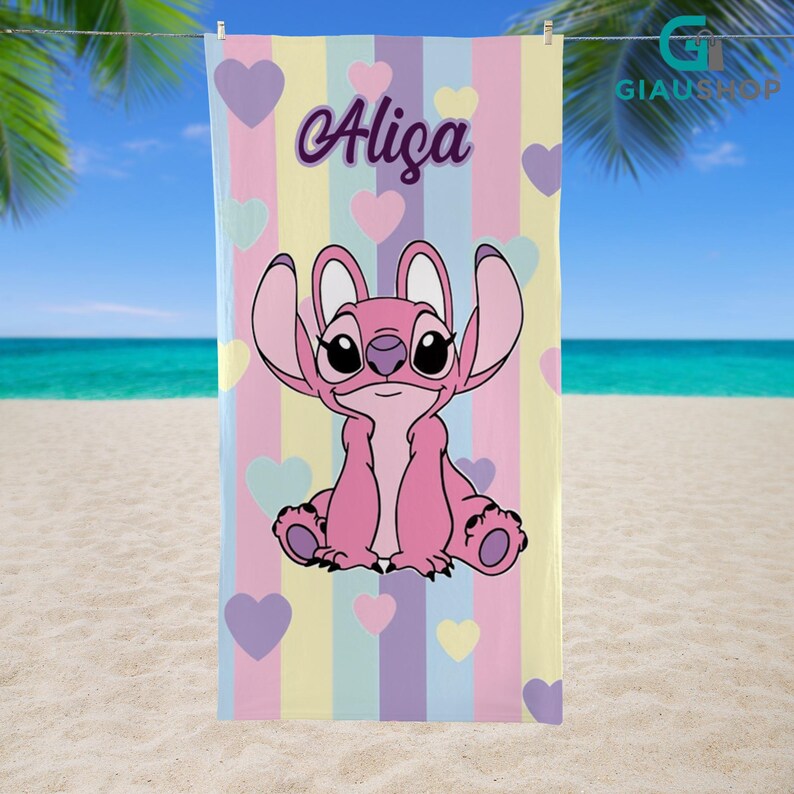 A colorful towel featuring a cartoon character, likely Stitch from the movie Lilo & Stitch, with the name "Aliza" printed on it. The towel is hanging on a line against a backdrop of palm trees and a beach.