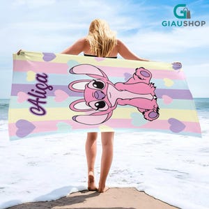 A woman holding a large, colorful towel with a cartoon character on it, standing on a beach with the ocean in the background.