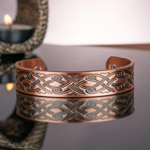 Puede incluir: Un brazalete de color cobre con un intrincado diseño de nudo celta. El brazalete es abierto en la parte inferior y tiene un acabado pulido. El diseño está grabado en el metal, creando un contraste. El brazalete se muestra sobre una superficie reflectante.