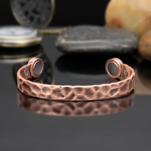 Può includere: Un bracciale a fascia color rame con una superficie martellata e testurizzata. Il design aperto presenta due inserti magnetici circolari a ciascuna estremità, con una finitura metallica e rientranze visibili lungo la fascia.