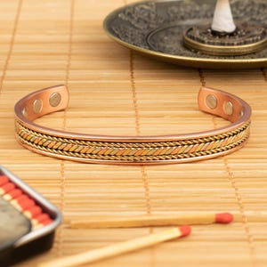 Op de afbeelding: Koperen armband met een gevlochten goud- en zilverontwerp. De open manchetarmband heeft twee magnetische sluitingen. De armband wordt gepresenteerd op een bamboemat.