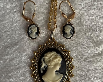 May include: A gold-toned cameo jewelry set featuring a pendant necklace and matching earrings. The pendant and earrings showcase a white profile against a black background, framed by ornate gold detailing. The set is displayed on a textured, white surface.