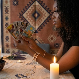 May include: A set of four tarot cards held in a fan shape, featuring detailed illustrations including a figure with a sun, a skeletal figure, and a star. The cards are rectangular with white borders and colorful, intricate artwork on the front faces.