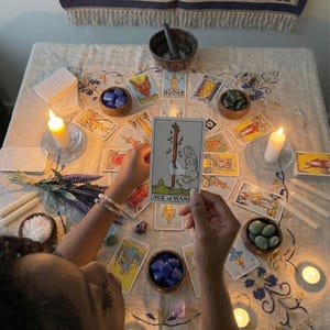 May include: A tarot card reading setup featuring an Ace of Wands card held in the center. The table is covered with a floral patterned cloth, surrounded by multiple tarot cards, small wooden bowls filled with crystals, lit candles, and lavender.
