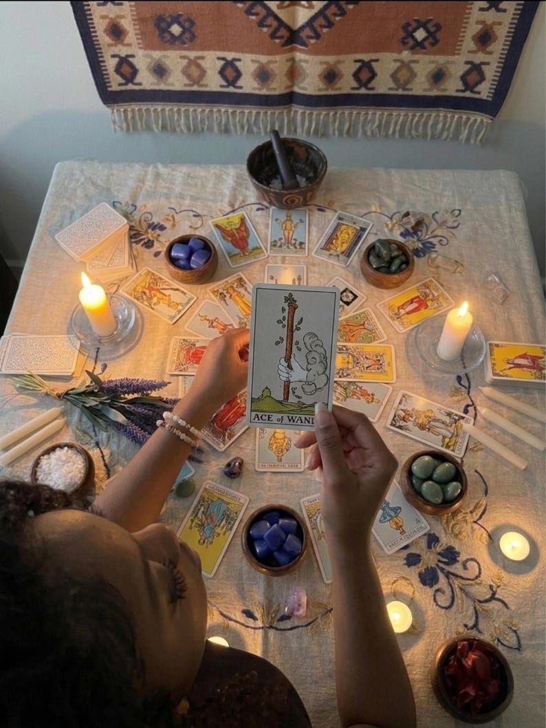 May include: A tarot card reading setup featuring an Ace of Wands card held in the center. The table is covered with a floral patterned cloth, surrounded by multiple tarot cards, small wooden bowls filled with crystals, lit candles, and lavender.