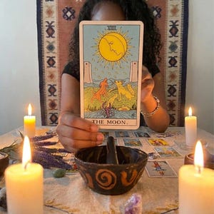 May include: A rectangular tarot card titled THE MOON featuring a yellow sun with a face, two dogs, a lobster, and two towers on a light blue background. The card is held upright above a dark ceramic bowl with spiral patterns and lit candles.