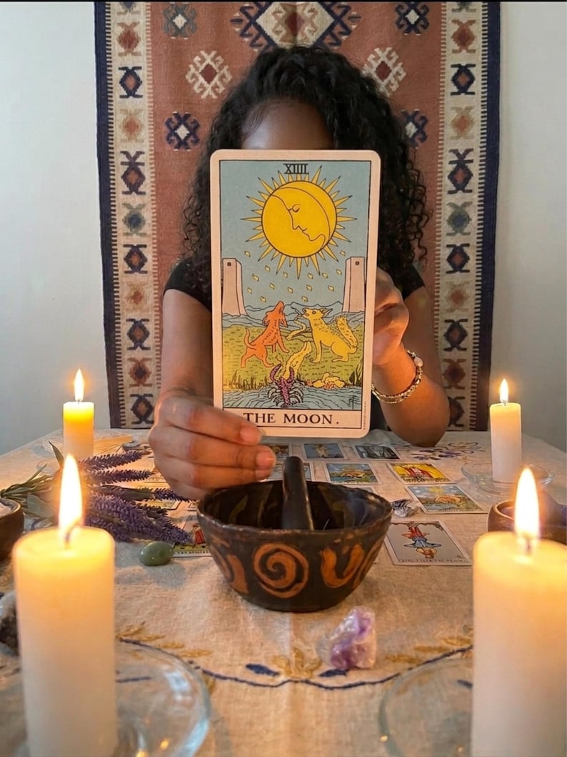May include: A rectangular tarot card titled THE MOON featuring a yellow sun with a face, two dogs, a lobster, and two towers on a light blue background. The card is held upright above a dark ceramic bowl with spiral patterns and lit candles.