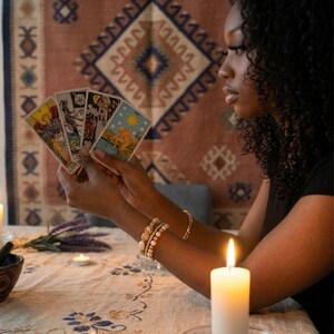 Peut inclure: Une personne tient des cartes de tarot, avec une bougie allum&eacute;e au premier plan. Les cartes pr&eacute;sentent des illustrations color&eacute;es. La sc&egrave;ne se d&eacute;roule sur une table avec une nappe &agrave; motifs floraux. Un tapis d&eacute;coratif est en arri&egrave;re-plan.