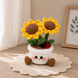 May include: A crocheted potted plant featuring two yellow sunflowers with brown centers, green leaves, and a small red ladybug. The white pot has a smiling face with pink cheeks and two brown feet, all crafted from yarn in a soft, textured style.