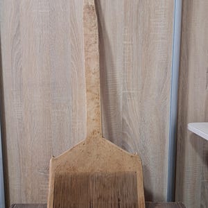 May include: A vintage wooden wool comb with a long handle and a wide, toothed head. The comb is a light brown color and appears to be well-worn, suggesting it has been used for many years. The handle is approximately 30 inches long.