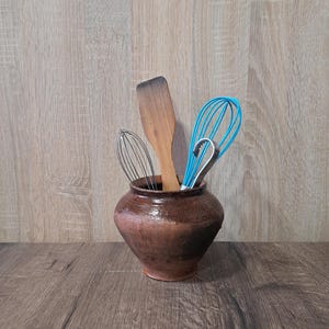 May include: A brown ceramic utensil pot containing a wooden spatula, a blue whisk, and a metal whisk. The pot is on a wooden surface, evoking a rustic kitchen aesthetic. The pot is approximately 15 cm tall.