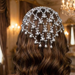Peut inclure: Un bijou de tête argenté avec un design en forme de filet, orné de nombreuses fausses perles de différentes tailles. Le bijou est porté sur des cheveux bruns ondulés, créant un look élégant et sophistiqué. Les perles sont en forme de goutte.