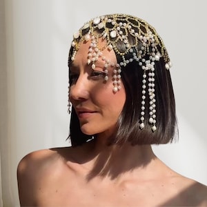 May include: An elaborate gold-coloured headpiece adorned with clear crystals and faux pearls. The headpiece, worn on the head, features an intricate design with strands of pearls cascading downwards. This elegant accessory is suitable for bridal or special events.
