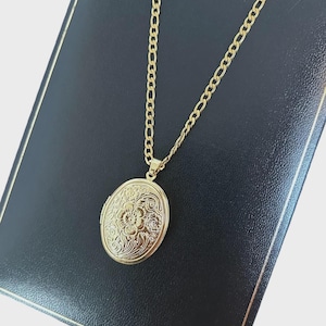 May include: A gold-coloured oval locket necklace with a floral design. The locket hangs from a gold chain. The necklace rests on a black surface.