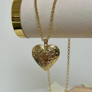 May include: A gold heart-shaped locket necklace with an intricate floral pattern. The locket is suspended from a fine chain. The necklace is presented on a white surface, with a gold bangle visible in the background.