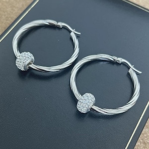 May include: A pair of silver hoop earrings. Each earring features a twisted design and a small, round charm set with clear crystals. The earrings are presented on a dark surface.