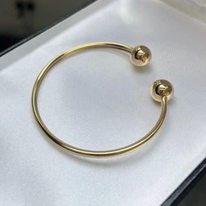 May include: A gold-coloured open cuff bracelet with a ball at each end. The bracelet is displayed on a white satin surface inside a black box. The bracelet's design is simple and elegant.
