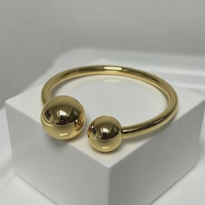 May include: A gold-toned open cuff bracelet featuring a smooth, polished finish. The bracelet is designed with a circular band that terminates at each end in a spherical ball, with one ball noticeably larger than the other, creating a balanced look.