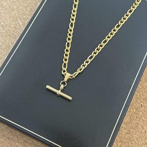 May include: A gold-coloured necklace with a T-bar clasp. The chain has a curb link design. The necklace is displayed on a black box with a gold border. The background is a light brown surface.