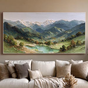 Puede incluir: Pintura al óleo de un paisaje montañoso con un lago. La obra presenta una paleta de verdes, azules y blancos, que representan colinas onduladas, picos nevados y una masa de agua serena. La pintura está enmarcada y colgada sobre un sofá blanco.