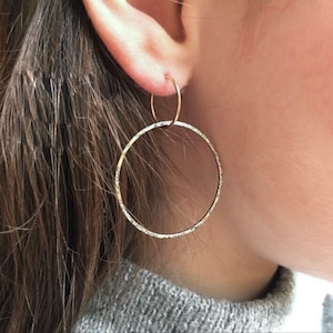 Puede incluir: Un par de pendientes de aro de plata con un acabado texturizado. Los pendientes se llevan en la oreja de una persona.