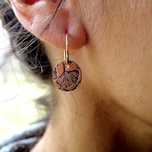 May include: Copper-colored round earring with a detailed ginkgo leaf design. The earring hangs from a gold-colored hoop. The ginkgo leaf is etched into the copper, creating a contrasting dark brown pattern. The earring is shown on a person's ear.
