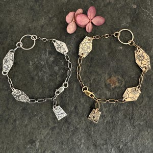 Pode incluir: Duas pulseiras com desenhos florais. Uma pulseira é de prata com um berloque de prata e a outra é de ouro com um berloque de ouro.