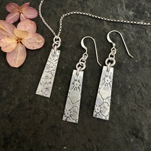 Op de afbeelding: Een zilveren ketting en bijpassende oorbellen met een bloemenmotief. De hanger en oorbellen zijn rechthoekig en hebben een delicaat bloemenpatroon.
