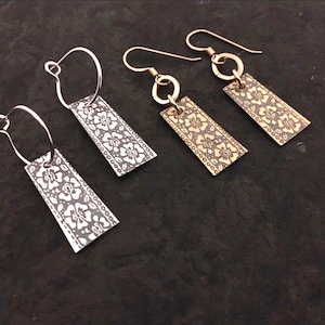 May include: Four pairs of rectangular earrings with floral patterns. Two pairs have silver-coloured hoops, and the other two have gold-coloured hooks. The earrings are displayed on a dark, textured surface.
