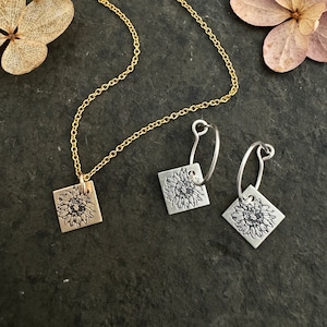 Pode incluir: Um conjunto de joias de ouro e prata com um design de girassol. O colar tem uma corrente delicada e um pingente quadrado com uma imagem de girassol. Os brincos são de argola com um pingente quadrado de girassol pendurado em cada argola.